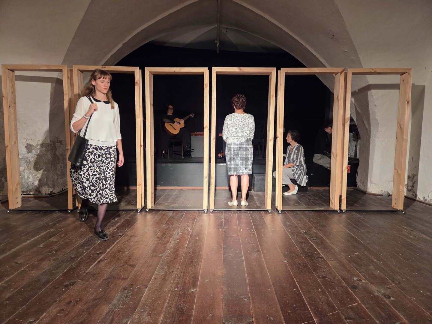 Theateraufführung mit vier Holzrahmen, Frau geht nach links, Gitarrist und zwei Frauen sitzen.