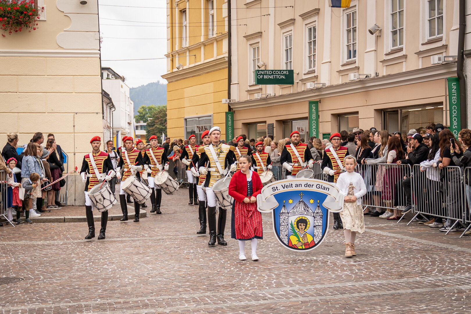 Marschierende Trommler in Uniformen von St. Veit an der Glan, begleitet von einem Mädchen, auf einer Gasse.